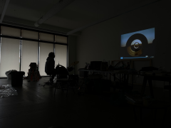 A person seen in profile sits on a stool in a dark room watching the projection of a film or video on the wall depicting the view through large cylindrical cement shapes with sky behind them.